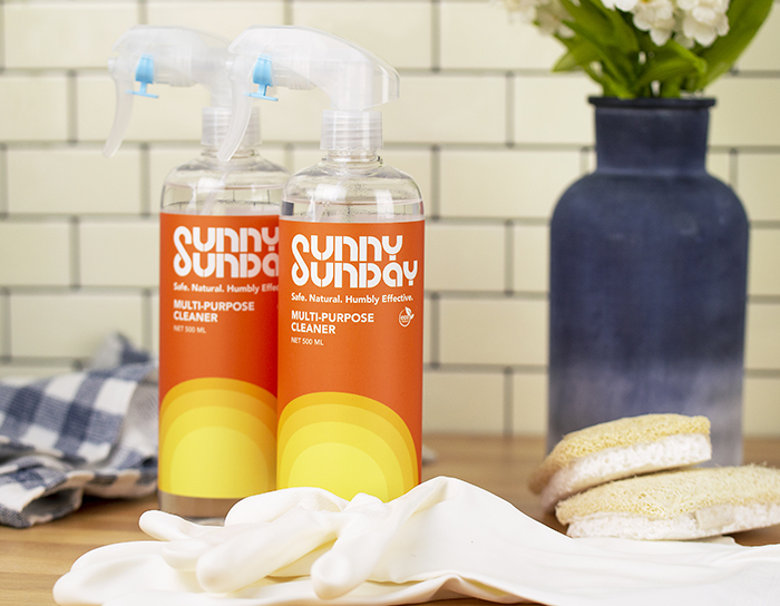 Luxury Spectacle® White Matte Roll Labels - Two bottles of 'Sunny Sunday' multi-purpose cleaner on a kitchen counter, with a backdrop of white subway tiles. The bottles have a vibrant gradient label from yellow to orange with a clear white text stating 'Safe, Natural, Humbly Effective', emphasizing the product's safe and natural cleaning properties. A blue vase with white flowers and cleaning supplies, such as gloves and sponges, are tastefully arranged around the bottles, evoking a clean, fresh environment.